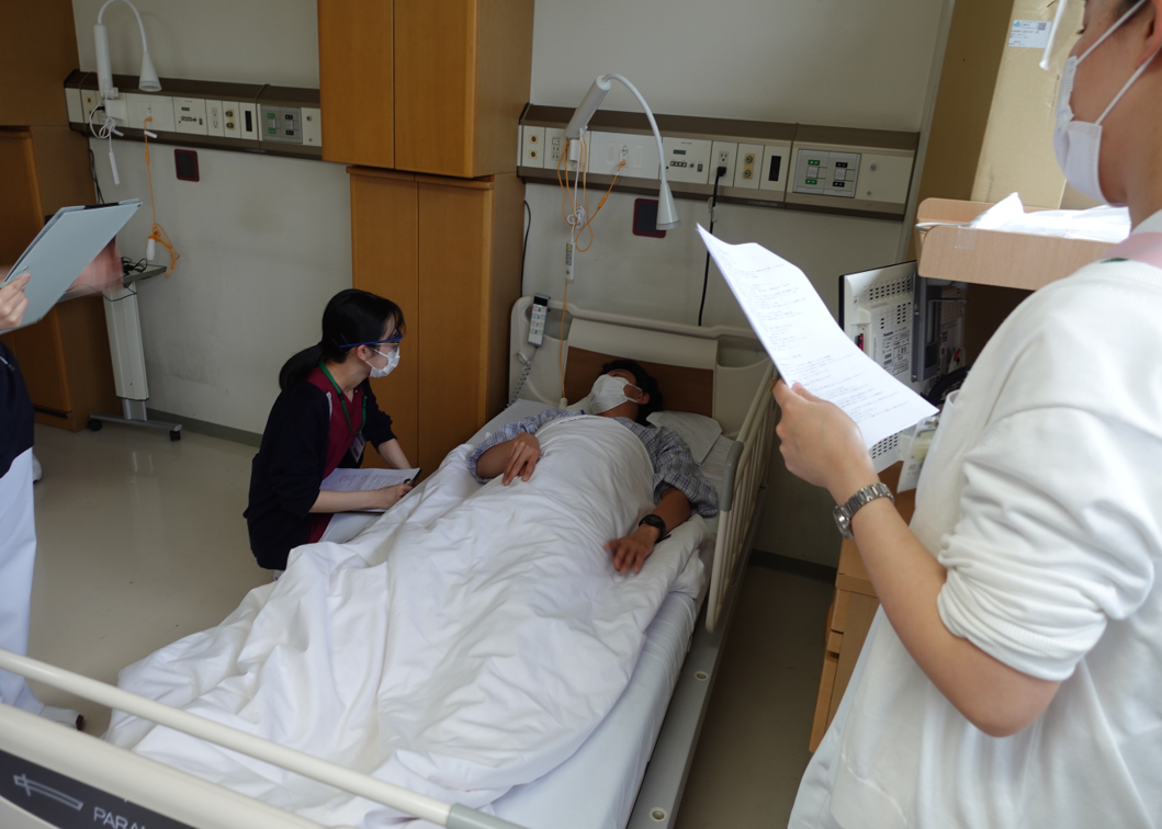 看護過程演習のようす。看護師が、ベッドに横になっている患者と目線をあわせて会話している。